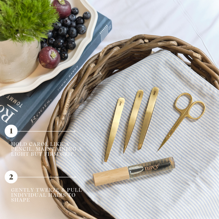 Set of gold tweezers and scissors on a textured surface with a basket and fruit in the background.