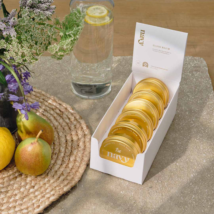 A white retail pack containing six gold-colored hand balm containers, displayed with an open lid and the brand name visible.