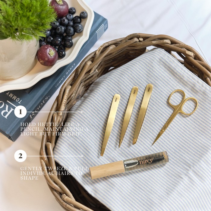 Set of gold tweezers and scissors on a textured surface with a basket and fruit in the background.