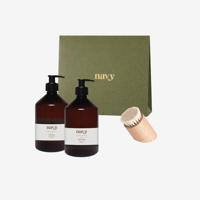A bath and body gift set by Navy, featuring two dark amber pump bottles with white labels, a sage green gift bag with gold lettering, and a natural wood scrubber with a white bristle head, all arranged on a light gray background.