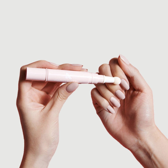 Hands applying Cuticle Serum Duo pen to moisturize nails with soft pink manicure on neutral background