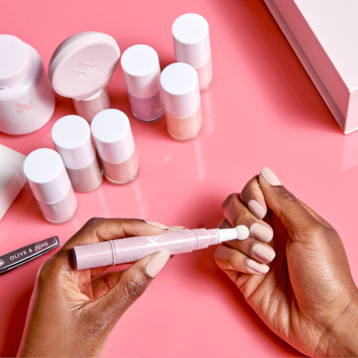 Hands applying Cuticle Serum Duo to moisturize cuticles on nails with soft beige manicure on pink surface