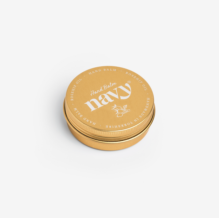 A gold-colored, round tin container with a label that reads 'Navy hand balm' and has some decorative floral design elements.