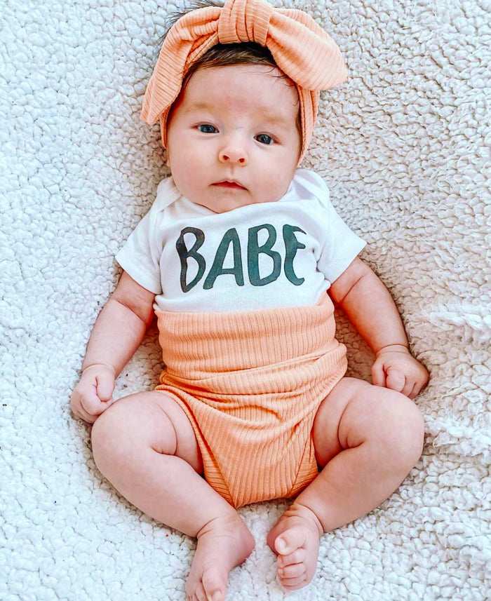 Baby wearing a 'Babe' shirt and peach outfit on a textured surface