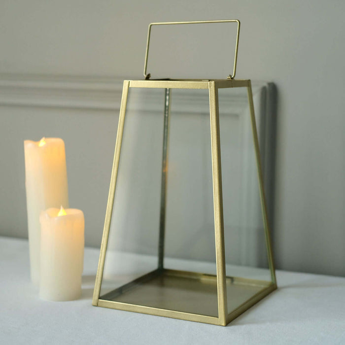 Gold geometric lantern with glass panels and handle beside lit candles.