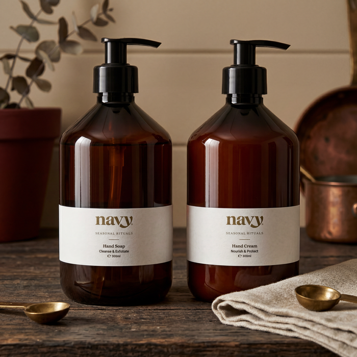 Two amber glass pump bottles labeled 'navy' Hand Soap and Hand Cream on a rustic wooden surface, with a folded beige cloth and small brass spoons nearby.