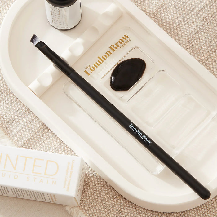 Eyebrow brush with black handle and white ferrule on a textured beige surface from the london brow company