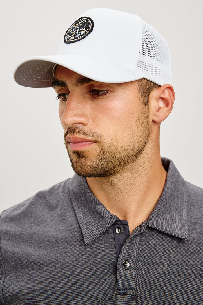 Golf Hat With Embroidered Branded Patch at Front, Adjustable Snapback, Mesh Back, and 6-Panel Construction in White Colorway - Upper Body Front View Featured on Model