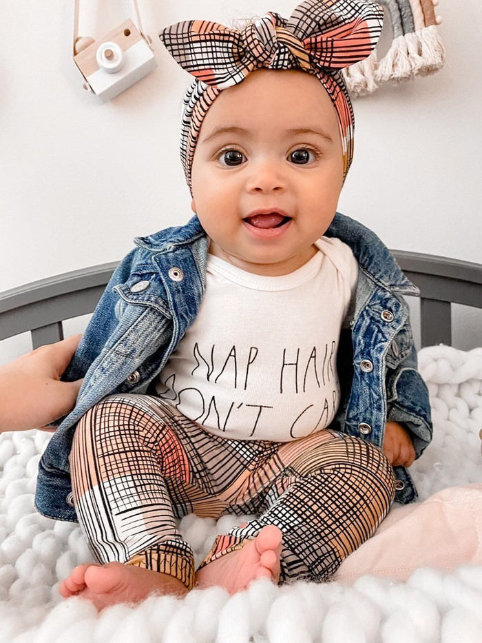 Adorable baby sitting in Retro Plaid Leggings paired with a matching plaid bow headband, denim jacket, and a 'Nap Hair Don't Care' onesie.