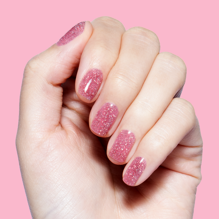 Hand with nails painted in Rosé Rhymes with Partay Gel showing cool-toned pink flash glitter gel polish mani on pink background