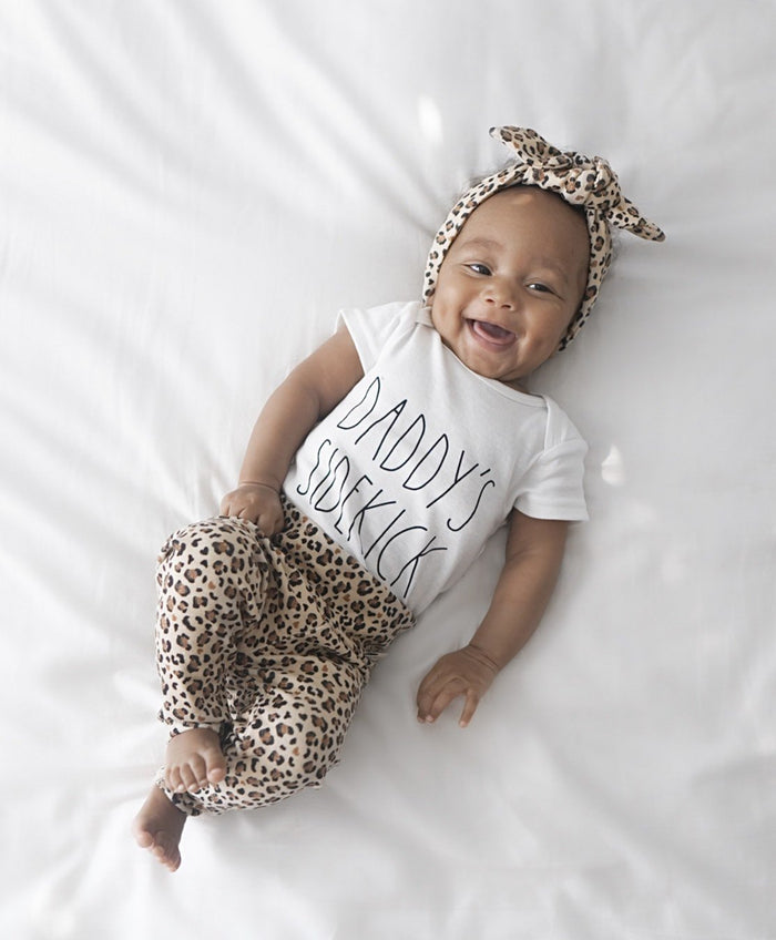 Smiling baby lying on a white blanket wearing leopard print leggings paired with a matching leopard headband and a "Daddy's Sidekick" white onesie.