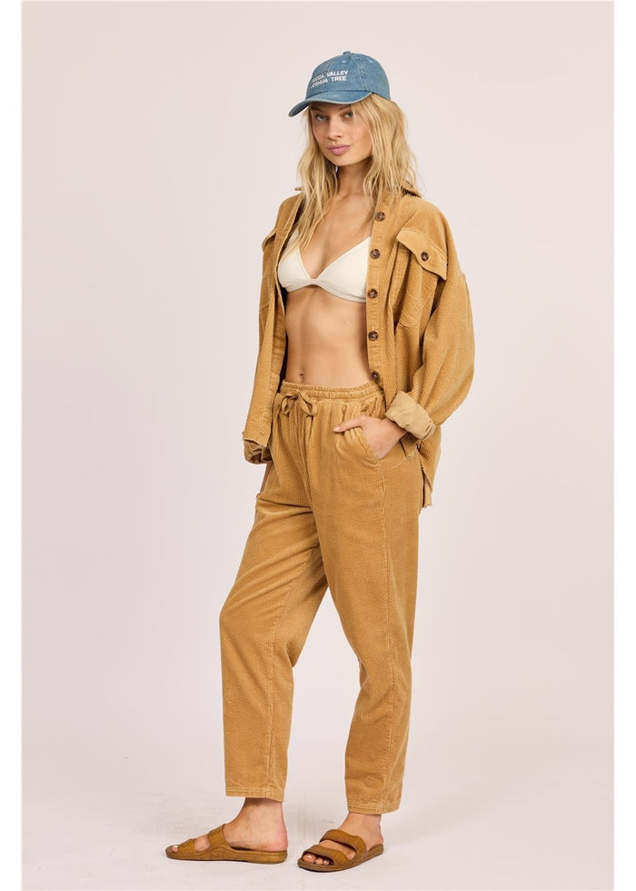 Woman wearing a beige jacket, pants, and cap with a white bikini underneath on a light pink background